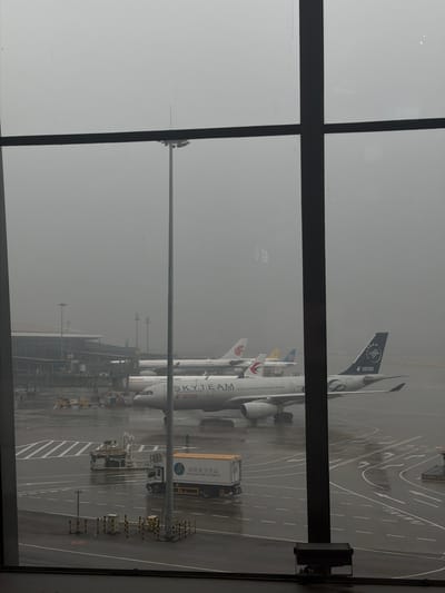 An overcast day at Guangyang District, China, likely at an airport, is depicted through a large window with dark framing. Several airplanes are parked on the tarmac, with one prominently featuring "SKYTEAM" branding. A delivery truck with "NANLAND AIR CATERING" written on its side is positioned near the planes. The tarmac appears wet, suggesting recent or ongoing precipitation. In the background, airport buildings and other aircraft are visible through the dense fog. The scene is static, with no discernible actions or interactions among individuals.