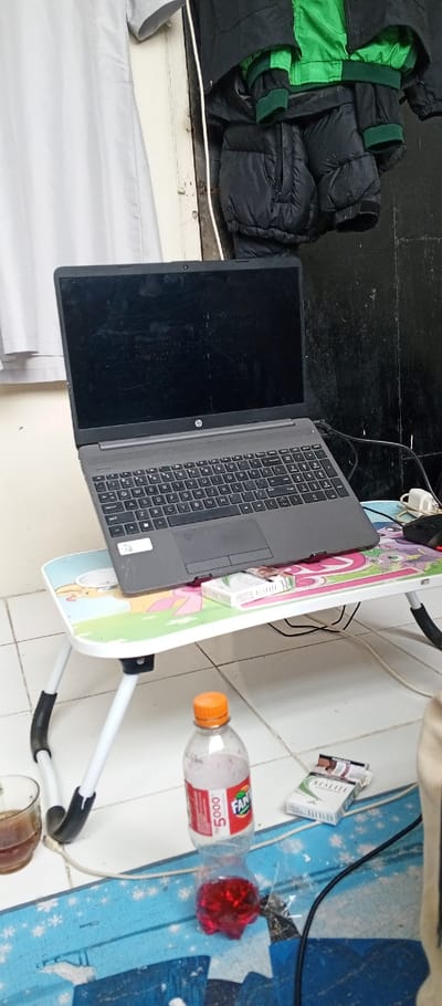 An indoor scene in Weintiapa, Liberia, depicts an unpopulated area with various personal items. In the mid-ground, a dark grey HP laptop with a dark screen is open and rests on a portable folding table featuring a multicolored, cartoon-themed surface. A white pack of "NESLITE" cigarettes is positioned on the table next to the laptop. The table has white, foldable legs with black rubber grips.

On the tiled floor in the foreground, a clear plastic bottle with an orange cap, displaying a "Fanta" label and "5.000", contains a red liquid at the bottom and a lighter, frothy liquid above it. To the left of the bottle, a clear glass contains a dark liquid. To the right, two cigarette packs are present: one white "NESLITE" pack and one partially opened brown-and-white "PENJUALAN" pack. Black and white cables are visible across the light-colored tiled floor, which has visible grout lines. A blue mat with white patterns is partially visible under the cables and bottle.

The background features a light grey fabric or curtain on the left, and a dark, possibly black, wall surface on the right. Suspended above the laptop, against the dark wall, are multiple pieces of clothing, including a green jacket with black trim and a black puffer jacket. A thin white cable runs vertically along the wall. No individuals are present in the image.