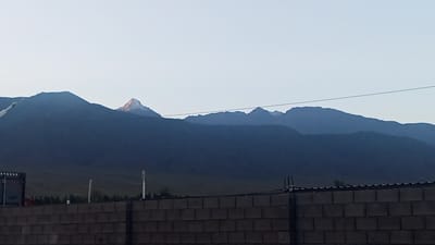 The image depicts a panoramic view of dark, silhouetted mountain ranges against a pale sky, likely at dawn or dusk. The mountains exhibit layered contours, with some peaks appearing snow-capped. In the foreground, a brick wall runs horizontally across the bottom of the frame, partially obscuring a landscape of sparse vegetation and utility poles. The lighting suggests an outdoor scene in a natural setting with man-made structures in the foreground. The location is identified as Choktal, Kyrgyzstan.