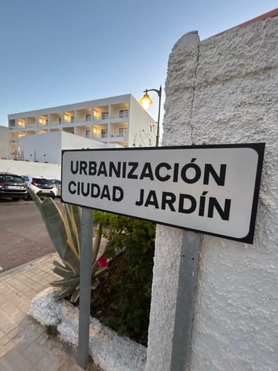 The image shows a street sign in Grao, Spain, that reads "URBANIZACIÓN CIUDAD JARDÍN." The sign is white with black lettering and is attached to a metal pole. The background features a white building with multiple stories and balconies, possibly a hotel or residential complex. A street lamp with a yellow light is visible above and to the left of the sign. To the left of the sign, there are several parked cars and a paved area. In the foreground, there is a plant with large green and yellow leaves, and a single pink flower. The environment appears to be an urban setting with a clear sky, suggesting it is daytime or dusk. The wall on the right side of the image is textured and painted white, with a visible crack.