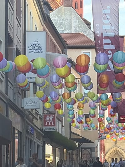 In Würzburg, Germany, on a street decorated with colorful paper lanterns, people walk by storefronts. The scene is captured from a slightly low angle, looking up towards the buildings and the lanterns strung across the street. Shops with signage like "Foto Weber" and "Walter Uhren & Schmuck" are visible. The architecture suggests an older European city, with buildings featuring multiple stories and traditional window designs. A large banner, partially visible, adds to the festive atmosphere. The overall impression is that of a vibrant street scene in a German city, likely during a festival or event.