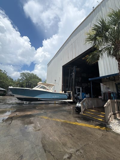 This media file shows a blue and white boat docked outside a large, corrugated metal building. The boat is positioned lengthwise along the building's opening, suggesting it is either being prepared for or has just been removed from storage. The building appears to be a workshop or storage facility, with various equipment and supplies visible inside, including what looks like an engine and shelving units.
The scene is set outdoors, with a bright, cloudy sky and lush green trees visible in the background. The ground is paved and appears wet in places, possibly indicating recent rain or cleaning. A yellow line is painted on the pavement, marking a boundary or pathway. A palm tree is prominent in the foreground, casting shadows.
There is some visible text on the boat: "Sea Queen" is written in cursive on the side, and a registration number appears to be "SC 1513 EH". Other text is visible on signs within the building, but it is too blurry to read.
The lighting suggests it is daytime, likely mid-morning or afternoon, given the brightness and the presence of shadows. The weather appears partly cloudy. The overall impression is of a working marine environment, possibly a boatyard or marina service center. No people are visible in the image, and the scene evokes a sense of quiet activity or readiness for a marine excursion. The location is identified as Hilton Head Island, United States of America.