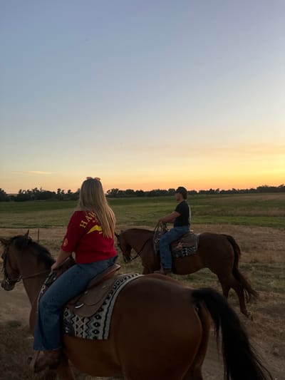 Two individuals are depicted horseback riding at sunset in an open field within Sloughhouse, United States. In the foreground, a blonde female, facing away from the camera, wears a red short-sleeved shirt with a yellow graphic on the back, blue jeans, and pink sunglasses perched on her head. She is mounted on a bay horse equipped with a Western saddle featuring a black, white, and tan patterned blanket. Ahead and to her right, a male rider, facing left, wears a dark short-sleeved shirt, blue jeans, and a dark hat. He is mounted on a second bay horse with similar Western tack, including a dark patterned saddle blanket. The horses are in a walking or standing posture on dry, grassy ground. The background features a distant treeline under a sky transitioning from pale blue overhead to orange and yellow at the horizon, indicative of late day.