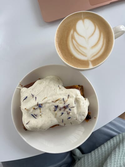 An overhead shot captures a coffee and cake arrangement on a white table in Callbach, Germany. A white ceramic mug filled with a latte featuring a detailed rosetta latte art pattern is positioned in the upper right. Below and to the left of the mug, a white ceramic plate holds a slice of cake, possibly carrot cake, topped with a substantial layer of white, speckled frosting. This frosting is garnished with scattered dried flower petals, primarily blue and light brown/orange hues. In the upper left corner, a portion of a rose-gold colored laptop is visible. The lower right section of the frame shows a light blue fabric item, likely clothing or a blanket. No individuals are visible.
