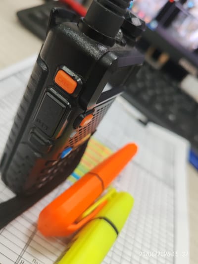 The image shows a close-up of a black walkie-talkie with an orange "CALL" button, resting on a document. Two highlighter pens, one orange and one yellow, are placed diagonally across the document, partially obscuring the walkie-talkie. The background is blurred, showing what appears to be a computer keyboard and monitor. A timestamp "21/06/2026 15:32" is visible in the bottom right corner. There are no people or animals in the image, and the scene appears to be indoors, possibly an office or study. The image does not contain any information that would allow for its location to be determined within Prikubanskii, Russia.