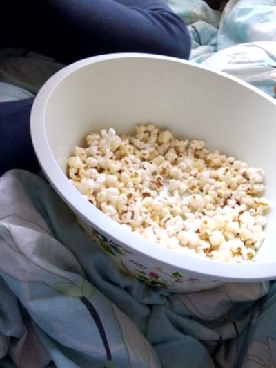 The scene depicts an indoor setting, reportedly in Kalimoni ward, Kenya. A white, oval-shaped bowl, subtly adorned with a muted floral pattern featuring green leaves and faint reddish elements on its lower exterior, is prominently featured in the center. The bowl contains a significant quantity of popcorn, primarily white and puffed, with some kernels exhibiting light yellow or slightly browned areas. The bowl rests upon or is partially surrounded by light blue textile material, which displays a repeating pattern of dark stems and light green, stylized leaves, consistent with bedding or a blanket. To the upper left, a segment of a human leg, covered in dark blue fabric, is visible, indicating the presence of an individual. A minimal portion of what appears to be a hand or fingers is discernible on the extreme right edge, near the bowl's rim. The posture and objects suggest the individual is in a relaxed state, likely consuming the popcorn.
