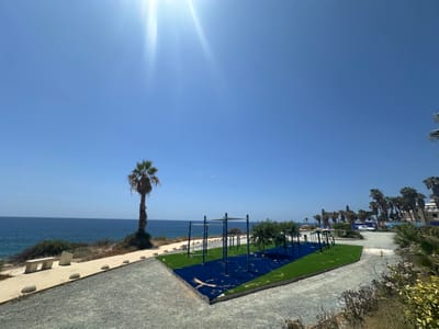 In Kissonerga Community, Cyprus, a bright, sunny day is captured with a clear blue sky and visible sun flare. The foreground features a gravel path leading towards a modern outdoor gym with blue rubber matting and green artificial turf. Several metal bars and structures make up the exercise equipment. To the left, a paved walkway runs alongside the sea, with a concrete bench and a lone palm tree. Beyond the gym, more palm trees are visible, along with buildings and a white vehicle on a dirt road. The ocean stretches out to the horizon, meeting the sky under the strong sunlight.