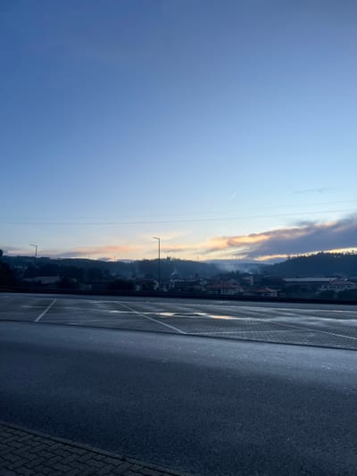 An angled outdoor shot depicts a parking lot meeting a distant town. The sky transitions from a deep blue to a lighter hue near the horizon, where the town sits nestled against hills. Smoke or mist rises above the rooftops, catching the colors of dawn. Utility poles are visible against the skyline. The parking lot is marked with white lines, and the pavement appears damp. A patterned border is visible in the bottom right corner.