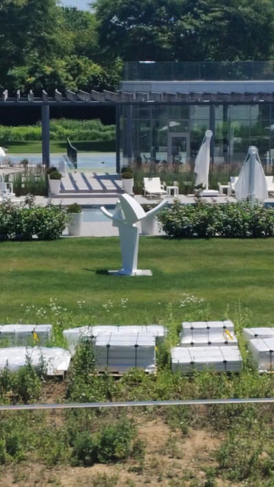 The image shows an outdoor scene featuring a modern building with large glass windows, a lush green lawn, and a patch of sandy, overgrown ground. Several white umbrellas are visible on a patio area adjacent to the building. In the foreground, a white abstract sculpture with curved elements stands on the lawn. The lawn is neatly mowed, with some small white flowers scattered across it.

To the right of the lawn, several stacks of rectangular objects wrapped in white plastic are arranged on wooden pallets. These appear to be construction materials. The background on the right shows a sloping area covered in dry grass and low-lying vegetation, suggesting a less manicured landscape.

A blue tennis court is visible in the mid-ground, beyond the patio area. The overall setting appears to be a resort or a well-maintained private property. The lighting suggests it is daytime, likely sunny, given the brightness and shadows. There is no visible text in the image. The scene is serene and devoid of people, focusing on the architectural and landscape elements. The image is captured from a slightly elevated perspective, looking down and across the scene.