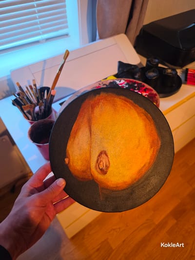 The image shows a close-up of a round canvas painting held by a person's hand, with a collection of paintbrushes in a red cup in the background. The painting depicts what appears to be a close-up of a breast. The background includes a white surface, possibly a desk or table, a window with blinds, and some art supplies. The lighting suggests an indoor environment, likely an art studio or a home. The presence of the artist's hand and the brushes indicates that the painting is either in progress or has recently been completed. The image does not provide any specific information about the location in Melhus, Norway.