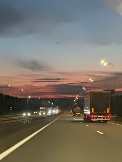 The image shows a highway at dusk. The sky is a gradient of blue and pink, with clouds casting shadows. There are several trucks on the road, including one in the foreground with a dark trailer and red taillights. Streetlights illuminate the highway. The road is paved and has white lane markings. A line of trees is visible on the left side of the road. The location is identified as the city of Zvanka, Russia.