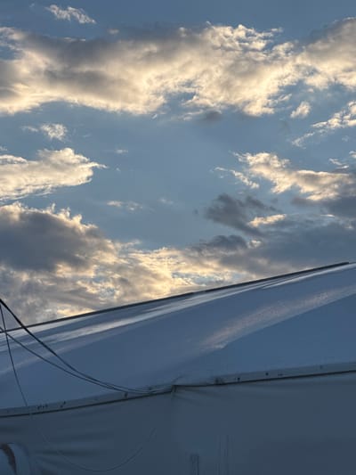 The image shows the roof of a white tent structure against a cloudy sky. The sky is a mix of blue and gray clouds, with patches of sunlight illuminating some of the clouds. There are some wires running across the top of the tent. The tent appears to be made of a white fabric material. The image is cropped, suggesting it is a close-up of a larger scene. The location is specified as Khadzhalmakhi, Russia, but there are no visible landmarks or identifying features in the image to confirm this location. There are no people or animals visible in the image.