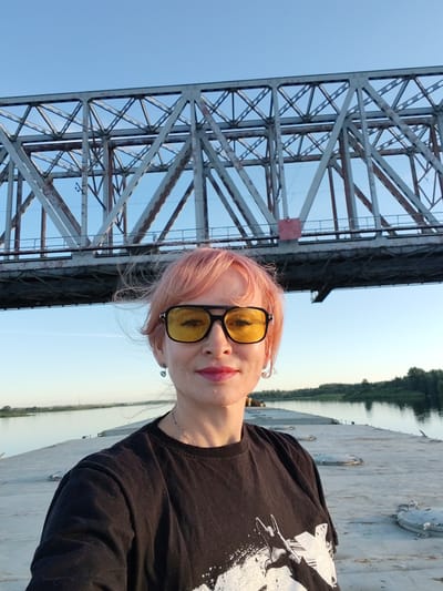 A woman with short, pink hair and yellow-tinted sunglasses takes a selfie on a barge. She is wearing a black t-shirt with a white graphic print on it. She has on dangling earrings and a necklace. Her lips are painted a vibrant pink. The barge is on a wide river, with a dense green forest lining the opposite bank.
Behind the woman, a large, grey metal railway bridge spans the river. The bridge is a complex structure of girders and supports, showing signs of age and rust in some areas. The sky is a clear, pale blue, indicating it is likely late afternoon or early evening, with the sun casting a warm light. The overall atmosphere is serene and peaceful, with the woman appearing relaxed and happy. The location is Tarkanovo, Russia, and the scene appears to be a calm summer day.