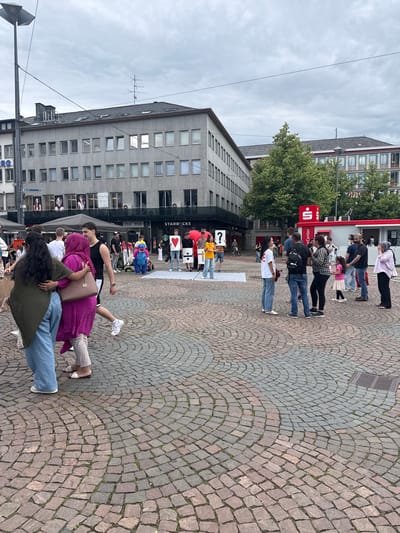 This image depicts a public square identified as Luisenplatz in Darmstadt, Germany, under an overcast sky. The square's surface is paved with a concentric pattern of brown and grey cobblestones, with sporadic green vegetation visible between the stones. A metal drain grate is present on the right side of the paved area.

Approximately 20-30 individuals are present in the scene, engaged in various activities. In the immediate foreground on the left, two women are interacting, one with dark hair wearing a dark green top and light blue jeans embracing another woman in a magenta dress, light-colored pants, and white sandals, who carries a light brown Michael Kors handbag.

Towards the mid-ground, a group appears to be participating in a public performance or demonstration on a large white sheet laid directly on the cobblestones. Four individuals stand on the sheet, holding large white signs: one displays a red heart, another a black question mark ("?"), a third a black division symbol ("÷"), and a fourth (partially obscured) also a red heart. Adjacent to this group, a person in a blue dinosaur costume with a yellow belly is standing.

To the right of the central performance, several smaller groups of people are standing and conversing. One group includes a woman in a white t-shirt and blue jeans facing a man in a black t-shirt with a black backpack. Nearby, a woman in a plaid shirt and black pants interacts with a man in a black t-shirt. A small child in a pink dress and white skirt is also visible within this cluster. Further right, a woman wearing a headscarf stands with a man in a black t-shirt.

In the background, commercial buildings line the square. A multi-story grey building with numerous rectangular windows is prominent on the left and center, featuring "Thieme" signage on the ground level and a partial "RG" sign. A "Starbucks" coffee shop sign is clearly visible. To the right, a red building section features the red square logo with a white "S" of Sparkasse bank, along with an "ec-center" sign and red banners. Dark canopy tents are situated near the buildings. Overhead power lines traverse the upper portion of the scene, and a large antenna structure is visible on the roof of a distant building. Several trees with green foliage are scattered throughout the background, particularly near the Sparkasse building. A tall lamppost stands on the far left.