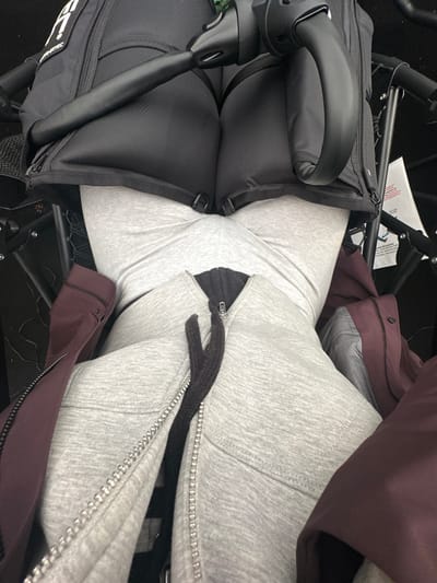 The image presents a first-person, downward view of an individual's lower torso and legs, situated in Weymouth, United Kingdom. The person is wearing a grey marl hooded sweatshirt, partially unzipped to reveal a dark inner layer, and light grey form-fitting leggings. A dark red or maroon jacket is draped over the individual's right side, partially obscuring the sweatshirt. In the upper portion of the frame, a black piece of equipment is prominently featured, comprising a smooth, curved black handle/bar and a textured black fabric component with visible stitching and multiple metal loops. The brand name "FANATEC" is partially visible on this fabric, alongside a stylized "A" or similar logo. A white product tag with red lettering is partially visible in the dark background on the right. The individual's body position suggests they are seated or standing in close proximity to the black equipment. The immediate environment is dark and appears to be an interior space.