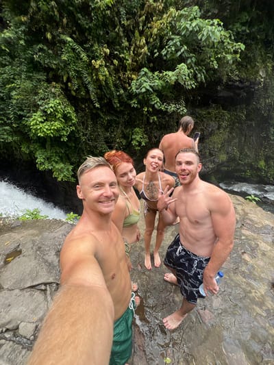 A group of five individuals, three males and two females, are positioned on wet, irregular grey and brown rock formations in Ambengan, Indonesia, with a lush green natural environment as the backdrop. The foreground male, with light-colored hair and bare-chested, wearing green swim shorts, extends his right arm, holding a device to capture a selfie. He smiles directly at the camera. To his right, a female with red hair styled in a bun, wearing a light green bikini, smiles slightly. Next to her, a female with light brown hair in a ponytail, wearing a white and brown patterned bikini top with a geometric chest tattoo, extends her left hand in a "V" gesture and protrudes her tongue. To her right, a bare-chested male with light brown hair, wearing black patterned swim shorts, smiles and holds a clear plastic water bottle in his left hand. All individuals in the foreground are barefoot. In the background, a fifth individual, a bare-chested male with light hair, is observed from behind, looking down at a mobile phone held in both hands. To the immediate left of the foreground group, a section of a white, turbulent waterfall or rapid is visible. The entire background consists of dense, verdant foliage covering a steep rock face, with a calmer section of a river flowing in the mid-ground on the right.