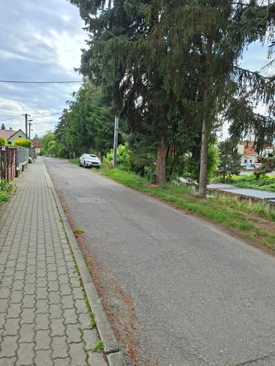 The image depicts a street scene in Ilava, Slovakia, on an overcast day. On the left, a paved sidewalk runs along a residential area with a metal fence and a house visible. The street itself is asphalt, with a white car parked on the right side. Tall trees line the right side of the road, with more trees and buildings in the background. A sign is visible on a building in the distance. The overall impression is of a quiet, suburban environment.
