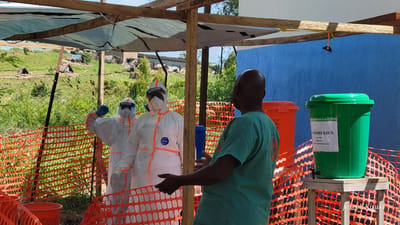This image depicts an outdoor health or hygiene checkpoint in Tchere, Democratic Republic of the Congo, under bright daylight and a clear sky.

In the foreground, a man in a teal medical uniform stands with his back to the viewer, gesturing with open hands towards two individuals. To his right, a green bucket serving as a handwashing station is placed on a wooden stand, with a spigot for dispensing liquid. An orange bucket is partially visible behind it. The area is enclosed by orange plastic safety netting, and a makeshift canopy of wood and mesh provides shade overhead.

The two individuals facing the man in teal are dressed in full white Personal Protective Equipment (PPE) suits, complete with face shields and blue gloves. One suit prominently displays the brand "Tyvek". Both face shields bear the text "Face Shield". One of these individuals has an arm raised, seemingly pointing or making a gesture towards the background.

The background shows a hilly, grassy landscape with scattered vegetation and a few simple, conical-roofed structures, possibly huts, behind a chain-link fence. The setting appears rural or semi-permanent.

Visible text includes "CHLORÉE 0,05%" on the label of the green bucket, followed by French instructions: "Ne pas boire" (Do not drink), "préparé le:" (prepared on:), and "toutes les 24 heures" (every 24 hours), suggesting the chlorinated water is for sanitation and needs daily replenishment. The orange bucket has "DES" written on its side. A "Joseph Kisuki" watermark is present in the lower right. The scene suggests an organized public health measure, possibly in response to an infectious disease outbreak.