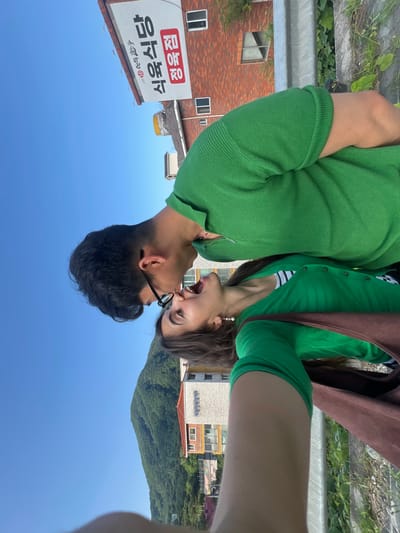 A couple is taking a selfie outdoors, with a man leaning in to kiss a woman. They are both wearing green tops. In the background, there is a sign with Korean writing, a brick building, a guardrail, greenery, a mountain covered in trees, and a clear blue sky. The image appears to be taken in Dongmyeong-myeon, South Korea.