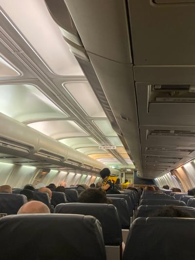The scene is the interior cabin of a commercial aircraft, presumably located at Athens International Airport "Eleftherios Venizelos" (ATH) within the Paiania Municipal Unit, Greece. The perspective is from a passenger seat, looking forward down the central aisle. Numerous passengers, primarily visible from the back of their heads and shoulders, occupy rows of dark blue/grey seats on both sides of the aisle. Approximately midway down the cabin, a person with dark hair, wearing a black top and an orange/yellow life vest, stands facing away from the camera, likely a flight attendant conducting a safety demonstration. Further forward in the aisle, another individual in a yellow garment is partially visible near the aircraft's front or galley area. The cabin ceiling features illuminated panels and overhead compartments, extending the length of the visible area. A red-on-white "EXIT" sign with a left-pointing arrow is illuminated above the aisle in the distance. Passenger service units are mounted below the overhead compartments.