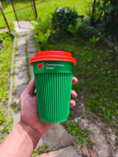 A person's hand holds a green disposable coffee cup with a red lid. The cup has a ribbed texture and features a white logo with the text "5 Пятёрочка Кафе" in Cyrillic script. The background is a slightly blurred outdoor scene, suggesting a natural setting with green foliage and a pathway. The lighting indicates it is daytime, likely sunny. The image seems to have been taken in a casual outdoor environment, possibly a backyard or garden, given the presence of plants and what appears to be a fence or gate in the upper left corner. The focus is on the coffee cup and the hand holding it, with the background providing context for the activity of enjoying a drink outdoors.