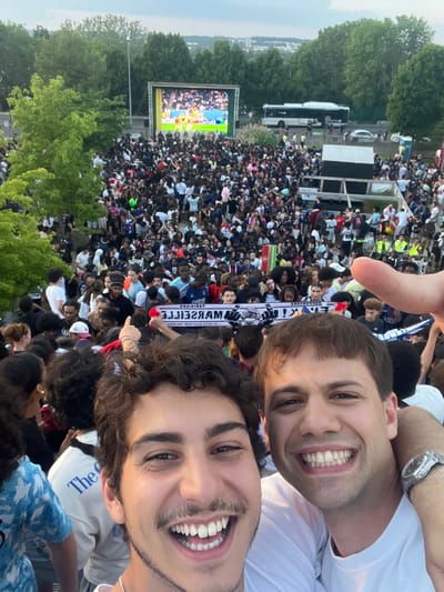 Two young men take a selfie while standing within a large crowd. The crowd is watching a broadcast on a large outdoor screen. There is a bus and a raised viewing platform adjacent to the crowd. The image was taken in Noisy-le-Grand, France.