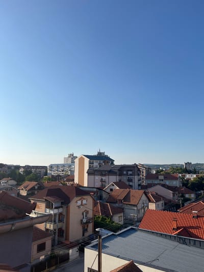This image captures a cityscape in Cacak, Serbia, under a clear, pale blue sky. The foreground is dominated by rooftops of various styles and materials, including red tile and corrugated metal. Beyond these, a densely packed urban landscape unfolds, featuring a mix of residential buildings and taller apartment blocks. Some structures are brightly lit by the sun, highlighting their architectural details and colors, while others are partially in shadow. Trees are interspersed throughout the city, adding touches of green to the urban fabric. In the distance, the landscape rises to gentle hills, indicating the city's geographical setting. The overall impression is one of a calm, early morning or late afternoon scene in a well-established urban area.