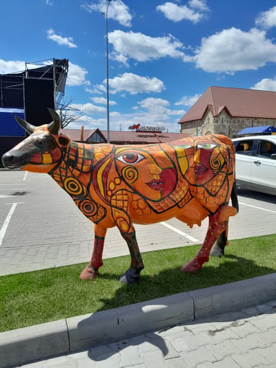 A vividly painted statue of a cow stands on a paved area next to a strip of green grass and a curb. The cow sculpture is adorned with a complex, colorful pattern of orange, yellow, red, and black. These designs include abstract shapes, eye-like motifs, and what appear to be stylized faces integrated into the cow's body. The cow is depicted with its head lowered and its legs positioned as if walking.

In the background, a building with a red tiled roof and stone walls is visible, suggesting an outdoor setting. A white car is parked to the right of the statue. The sky above is a bright blue, dotted with fluffy white clouds, indicating fair weather and likely daytime. To the left, there is a metal structure with scaffolding and black fabric, possibly a stage or event setup, against the sky. A streetlamp is also visible.

The overall scene appears to be an outdoor public space, perhaps a town square or an area for an event. The sunlight casts shadows, suggesting it is mid-day. No people are clearly visible in the frame, but the shadows indicate movement or the presence of unseen individuals. The artwork on the cow is the most prominent and detailed element. The location is identified as Velykyi Khodachkiv, Ukraine. No discernible text is visible on the statue itself, but a sign with Cyrillic lettering is partially visible in the background.