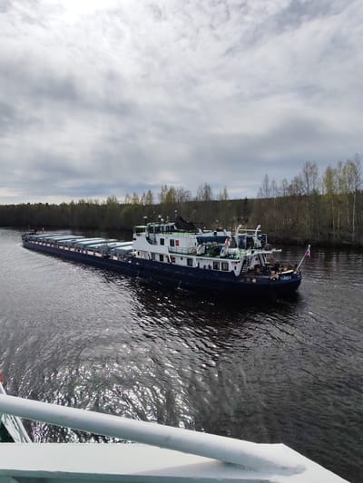 A long, blue and white river barge named "Павел" (Pavel) is moving along a waterway in Srednii Rubezh, Russia. The barge is heavily loaded with rectangular, gray containers. The body of water shows rippling reflections of the sky and surrounding trees. Bare trees line the riverbank, and a cloudy sky dominates the upper part of the frame. A portion of a white railing is visible in the foreground, likely from another vessel.