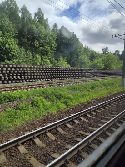 The image, taken from a moving train, shows a railway track running through a green, overgrown landscape near Ashukino, Russia. Parallel to the main track, there is a second track with a large stack of precast concrete elements alongside it, likely intended for rail construction or maintenance. A dense line of trees borders the tracks, and overhead power lines are visible against a cloudy sky. The view is slightly obscured by raindrops on the window.