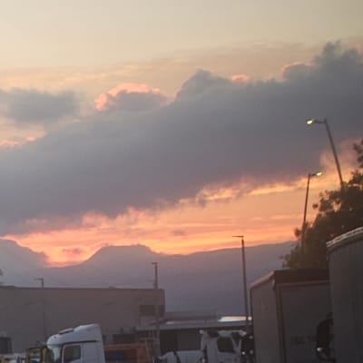 This is a photograph of a sunset over the city of Gurb, Spain. The sky is a gradient of orange, pink, and purple, with large, dark clouds obscuring parts of the horizon. In the foreground, there are several large trucks, some with their trailers visible. There are also streetlights and some trees. The overall impression is one of a dramatic and colorful sunset over an industrial or commercial area.