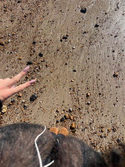 The image depicts a top-down view of a pebbly and sandy beach in Worthing, United Kingdom, under bright natural light. The ground consists of wet, light brown sand intermingled with numerous small, multi-colored pebbles and stones, showing reflections from sunlight. The lower portion of the frame shows the upper thighs and feet of an individual. The individual's feet are clad in light brown, Ugg-style boots. A dark, textured garment, possibly a coat or jacket, covers the legs and lower torso, with a white cord (likely a headphone cable or drawstring) visible against it. A left hand, featuring long, light pink artificial nails, extends into the upper-left quadrant of the frame, performing a 'peace' sign with the index and middle fingers extended.