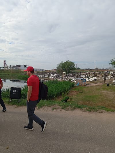 The image depicts a scene in Sol-Iletsk, Russia, specifically in an area that appears to be a recreational or resort zone. In the foreground, a person wearing a red cap and a red t-shirt is walking away from the camera, carrying a black backpack. They are wearing grey pants and black sneakers. To the left, the legs of another person are visible, also walking.

The environment features a body of water, possibly a lake or river, with reflections on its surface. Adjacent to the water is lush green grass and reeds. Further into the background, there is a large expanse of beach chairs and umbrellas, suggesting a public beach or resort. In the distance, buildings and structures are visible, including what looks like a mine shaft headframe on the left side. The sky is overcast with clouds. A black trash bin with a smiley face graphic is positioned near the path. The overall scene suggests a day at a leisure facility in Sol-Iletsk.