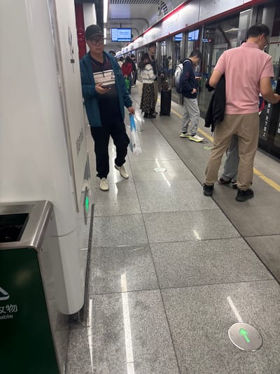 A subway platform with waiting passengers. The floor is gray tile with a green directional arrow. People are near the train doors. One man stands near the camera, wearing a blue jacket and black pants, looking at his phone. Others are further down the platform, including one wearing a pink shirt and khaki pants. A green trash receptacle is in the bottom right corner.