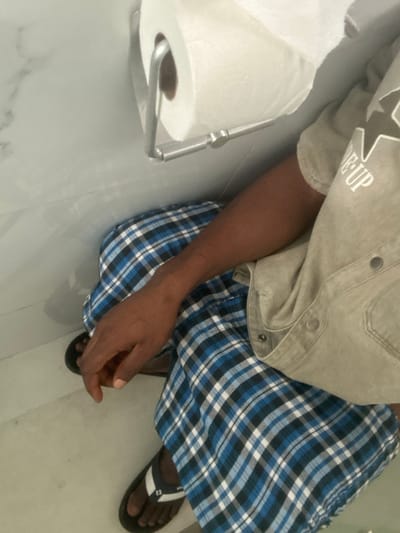 This image, captured in Otampa, Nigeria, presents an individual from a high-angle perspective within what appears to be a tiled bathroom or toilet area. The background features white ceramic tiles on the wall, where a roll of white toilet paper is mounted on a silver-colored metal holder. The subject, exhibiting dark skin, is partially visible, primarily showing their torso and lower body. They are wearing a light beige short-sleeved shirt with silver snap buttons down the front and the partial text "THE UP" visible on the upper left chest/shoulder area. Their lower body is clad in blue, white, and black plaid pants. Both feet are visible, shod in black flip-flops with a white stripe. The individual's right forearm and hand rest upon their plaid-covered lap, suggesting a seated or squatted posture on a light-colored floor.