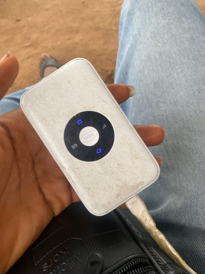 The image depicts a close-up, downward-angled view of a person holding a white, rectangular portable Wi-Fi device (MiFi) in Iyare, Nigeria. The device, notably scratched and smudged, features a prominent black circular interface with a white "MTN" logo at its center. Surrounding the logo, four blue illuminated icons are visible: a message envelope, a signal strength indicator with three bars, a battery level indicator, and an icon resembling a globe or a power button. A white charging cable, visibly reinforced with clear tape near the connector, extends from the bottom edge of the device, connecting downward out of frame. The person's dark-skinned left hand, featuring long fingernails, is holding the device. Below the device and hand, a black textured bag, partially revealing "MICHAEL KORS" text in white, is visible. The person's leg, clad in light blue denim jeans, occupies the right side of the frame. In the blurry background, a foot wearing a grey sandal rests on light brown, dusty ground.