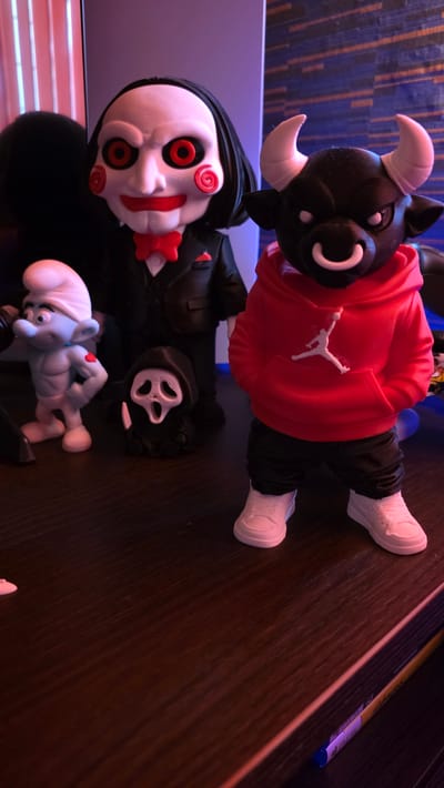 The image shows a collection of figurines on a dark wooden surface. Included are a black bull figure wearing a red hooded sweatshirt, a figure with a white mask featuring red eyes and a red bow tie, a black ghost-like figure with a white mask, and a blue Smurf-like figure. A pen or marker is visible on the right side of the image. The location information is not discernible from the image.