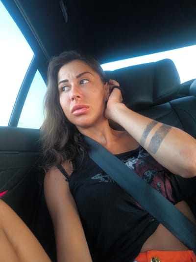 A woman is seen from the chest up, seated in the back of a vehicle. She has long, dark hair and is looking to her left. She is wearing a black tank top with a red and black graphic design and an orange skirt with gold accents. A black seatbelt is across her chest. Her left arm is raised to her head, revealing two parallel, horizontal tattoos on her forearm. The background consists of a black car interior and a bright sky visible through the car window. The image is a medium shot, taken from a slightly elevated angle. The location is identified as Municipal Unit of Assiros, Greece.