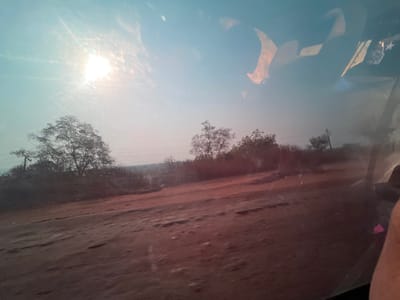 An outdoor scene with a dirt road in the foreground and trees in the background. The sky is bright with the sun. Telephone poles are visible. The image appears to be taken through a window, with reflections visible. The landscape is somewhat arid, with sparse vegetation.