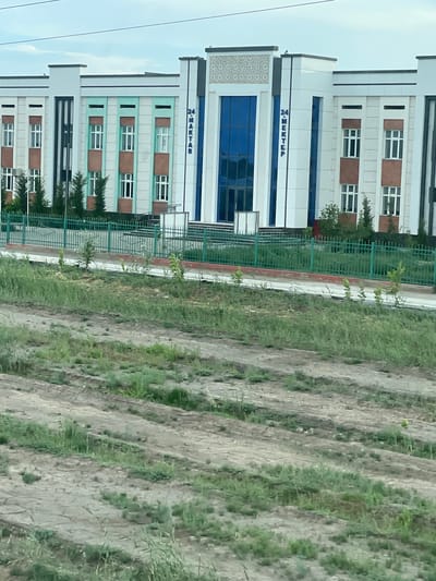 This is a vertical, outdoor photograph of a modern, multi-story building that appears to be a school or administrative complex. The building has a white facade with alternating bands of light green and brown panels. It features numerous windows, some with white frames and others with dark blue glass. The building is bordered by a green metal fence. To the right of the building, there is a patch of land with uneven terrain, covered in sparse grass and dry soil. The sky is visible in the upper left portion of the image and appears overcast.
There is text visible on the building in blue lettering. The first line, on a white section above a blue-glass window, reads "MEKTEB". Below this, on a green band, another word, also in blue lettering, appears to read "2-MEKTEB".
The scene suggests a daytime setting, possibly in the late morning or early afternoon, given the ambient light. The weather appears to be fair, with overcast skies. The presence of the building and the surrounding undeveloped land hints at a location in a developing or suburban area. No people or specific events are depicted. The overall impression is one of institutional architecture set against a backdrop of open land.