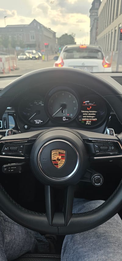 This media file shows the interior of a Porsche, with a focus on the steering wheel and dashboard. The driver's legs, clad in denim, are visible on the right side of the frame, suggesting the perspective of someone seated in the driver's seat. The steering wheel prominently features the Porsche logo. The dashboard displays various readouts, including a tachometer, speedometer, and a digital display showing "45779" and "2321," likely representing mileage and trip distance. A message on the digital display in German reads: "Fahrzeug Verkehrs- sicher abstellen und Anhänger- Kupplung verriegeln," which translates to "Park vehicle safely and lock trailer hitch." Other visible information includes "359 km," "18:43," and "24.0," possibly indicating range, time, and temperature. A "DRIVE MODE" selector is also visible on the steering column.

The scene appears to be captured during daylight, with natural light filtering in from outside. The view through the windshield reveals a street scene with other vehicles, suggesting the car is either in traffic or parked on a street. The presence of traffic lights and buildings in the background hint at an urban or suburban setting. The overall impression is that of a functional vehicle interior, with the driver possibly about to embark on a journey or having just completed one. There are no explicit emotional cues, but the focus on the car's controls and display suggests an interest in the vehicle's operation. The location context of Verviers, Belgium, provides a geographical reference for the setting.