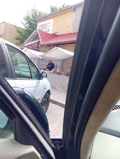 The image shows a street scene in Kuršėnai, Lithuania. In the foreground, the interior of a dark car is visible, with the open door and window of another vehicle taking up a significant portion of the frame. Through the open door, a light-colored car is parked on a paved surface. Beyond the car, there are market stalls with white awnings set up in front of a building. One stall has a banner that reads "Radviliškių KEPY" and another stall has a banner that says "MANO SAMAS". A man in a dark blue jacket and a light-colored hat is standing behind a stall, appearing to interact with customers or arrange goods. There are loaves of bread displayed on the tables. The building behind the market stalls is light yellow with a red-tiled roof and a sign that says "CSSS ŠIAULIAI". Trees are visible in the background, and the sky is overcast. The overall scene suggests an outdoor market or fair in a town.