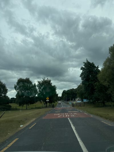 The image shows a view from a vehicle traveling on a road in Curragh Edge, Ireland. The road has a red marking indicating a "SCHOOL ZONE" with "SLOW DOWN" painted in white letters. A white line divides the lanes, and yellow lines mark the edges of the road. The surrounding environment is rural, with green grassy fields, fences, and numerous trees on both sides of the road. The sky is overcast with dark, heavy clouds, suggesting a cloudy or rainy day. In the distance, a few vehicles are visible on the road, and some buildings are present on the right side. There is a yellow triangular warning sign on a pole to the right of the road.