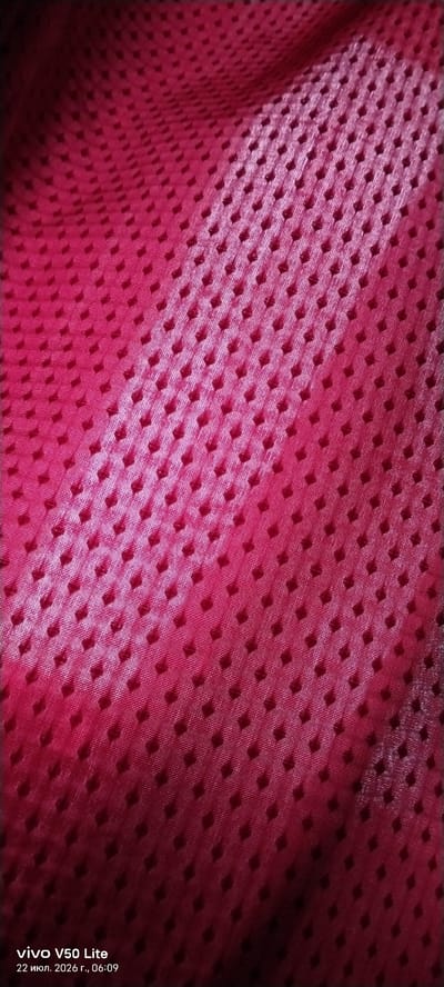 The image is a close-up, high-angle, full-frame shot of a textured, deep pink fabric. The fabric has a subtle sheen and is arranged in soft folds, creating shadows and highlights that emphasize its texture. The texture consists of a repeating pattern of small, raised diamond-shaped indentations. The lighting is dim, creating a gradient of light and shadow across the fabric, with the top left corner being darker and the lower right corner being brighter.
The bottom left corner of the image displays text which reads "vivo V50 Lite" and "22 июл. 2026 г., 06:09". This text indicates that the image was captured by a vivo V50 Lite phone on July 22, 2026, at 06:09. This timestamp is a data point associated with the image file.
The location in the city of Imeni Sverdlova, Russia is not depicted or implied by the image itself. The image solely contains a visual representation of fabric texture.