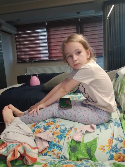 In an indoor residential setting, a young girl with blonde hair sits on a bed with a floral comforter. She is wearing a light-colored t-shirt with a graphic design and light purple pajama pants adorned with butterflies. Her left arm rests on her lap, and her right arm is extended across her legs. A smartphone is visible on her lap. To her left, a pile of colorful clothes includes a striped towel and a grey garment. A pink sock is placed on the comforter to her right. In the background, a dark blue sofa is visible, with a pink, kettle-shaped object and a pillow on it. The room features blinds on a window, a radiator, and an air conditioning unit. The scene appears to be within a city in Ukraine, likely Lobacheve, based on the provided information.