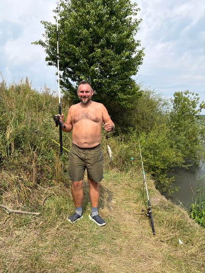 A shirtless man with a beard stands on a grassy embankment next to a body of water in Ruda-Siletska, Ukraine. He is holding a fishing rod in his left hand and a small fish in his right. He is wearing green shorts and grey socks with dark shoes. Two other fishing rods are planted in the ground to his right. Behind him are dense green bushes and trees, with a cloudy sky above. The ground is covered with dry grass and some scattered sticks.