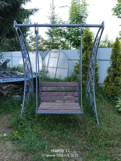An outdoor scene in Космос-2 Снт, Russia, features a weathered, grey metal garden swing with a wooden slatted seat, positioned on a grassy area dotted with wildflowers. Behind the swing, a pale blue fence separates the foreground from a backdrop of various trees, including evergreen bushes and taller conifers. To the left, a pile of discarded wooden planks and a green tarp with lettering are visible. Two large, clear geodesic dome structures are situated behind the fence. The sky above is overcast. A watermark at the bottom right corner of the image reads "realme 12 21 июня 2026 г. в 20:15". There are no people or animals present in the image.