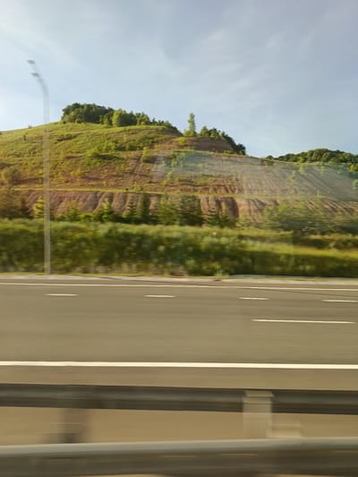 The image depicts a highway scene with a forested hill in the background. The highway has distinct white lane markings, indicating it is a multi-lane road. A silver guardrail is visible along the side of the road, separating the lanes from the embankment. The hill is covered with green vegetation and appears to be terraced in some areas, suggesting agricultural use or landscaping. Trees are scattered across the hillside, with a denser cluster at the crest. The sky is a light blue with some wispy clouds. The image is blurred due to motion, suggesting it was taken from a moving vehicle. The location is identified as Isakovo, Russia.