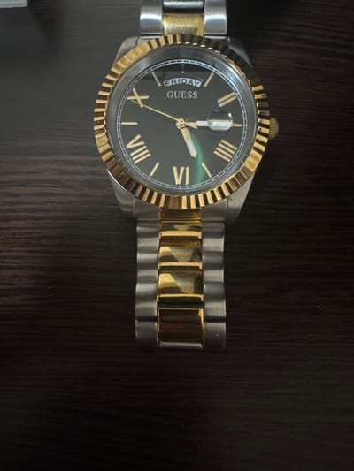 The image shows a close-up of a two-tone (silver and gold) Guess watch with a black dial. The watch has Roman numerals as hour markers and a day and date window at the 3 o'clock position, displaying "FRIDAY" and "18". The bezel is gold and fluted. The watch is placed on a dark, wooden surface, likely a table or shelf, with vertical wood grain visible. The lighting is dim, suggesting an indoor setting. No people are visible in the image.