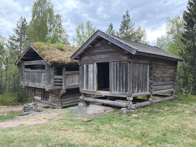 The image depicts two traditional Norwegian log buildings, likely located in an open-air museum or historical site. The building on the left features a sod roof covered in vegetation. The building on the right has a conventional wooden roof. Both structures are elevated on support beams and stones. The surrounding environment consists of grassy terrain and trees, with an overcast sky. The specific location in Drammen, Norway is not directly identifiable from the image.